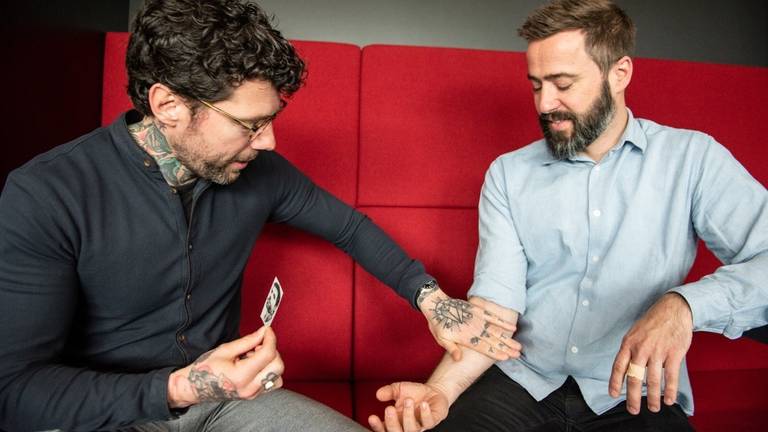 Henk van Straten plakt tatoeage met zelfportret op arm van boekhandelaar Jan Verhagen (Foto's: Cloakture photography)